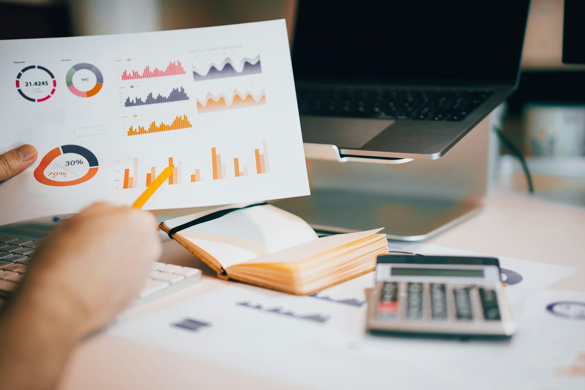 An analyst reviewing printed charts beside a laptop, notebook, and calculator